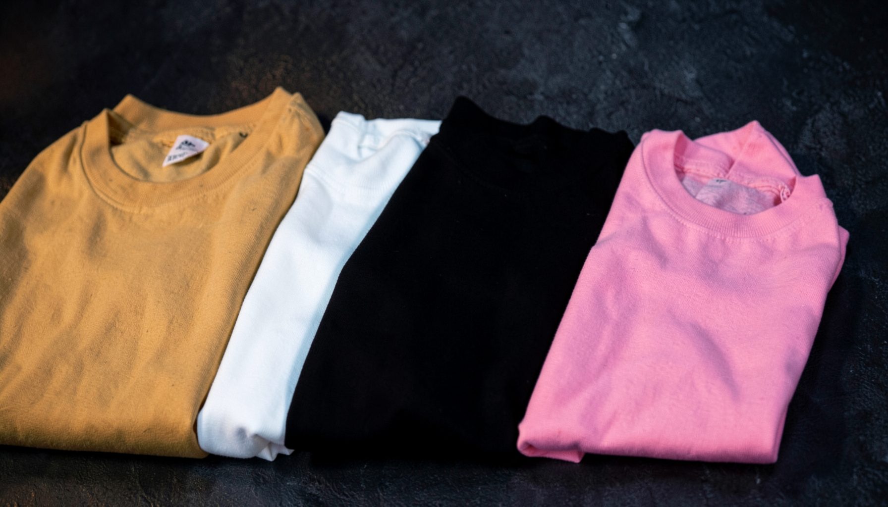 A row of folded t-shirts in tan, white, black, and pink against a dark textured backdrop, softly lit with warm and cool tones