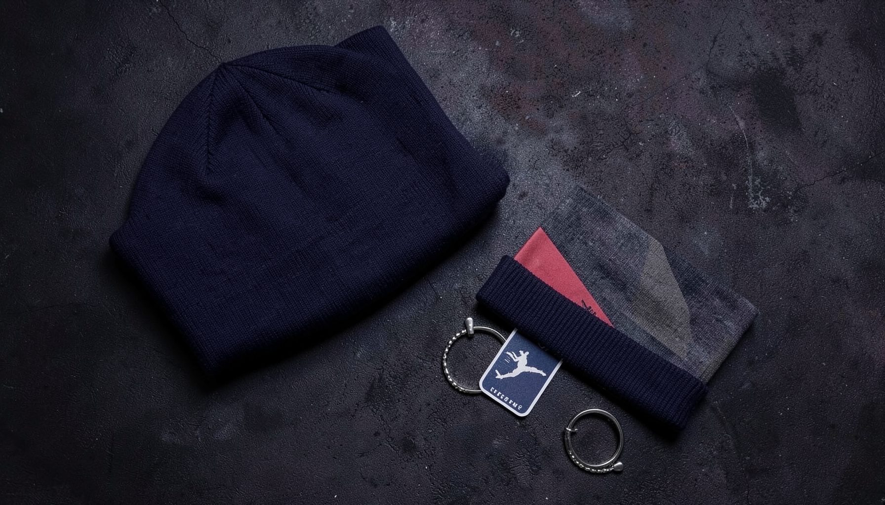 A flat lay of streetwear accessories on a dark textured surface — a beanie, keychain, and sticker arranged casually