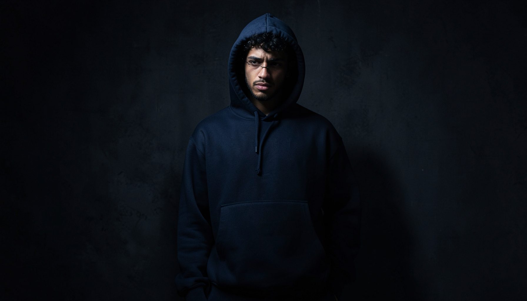 Dark moody streetwear scene with a hooded sweatshirt in soft shadow, deep navy and charcoal tones
